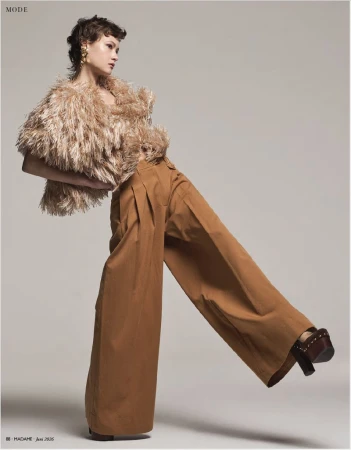 Model in a textured fringed top and wide-leg brown trousers posing against a neutral grey studio background.