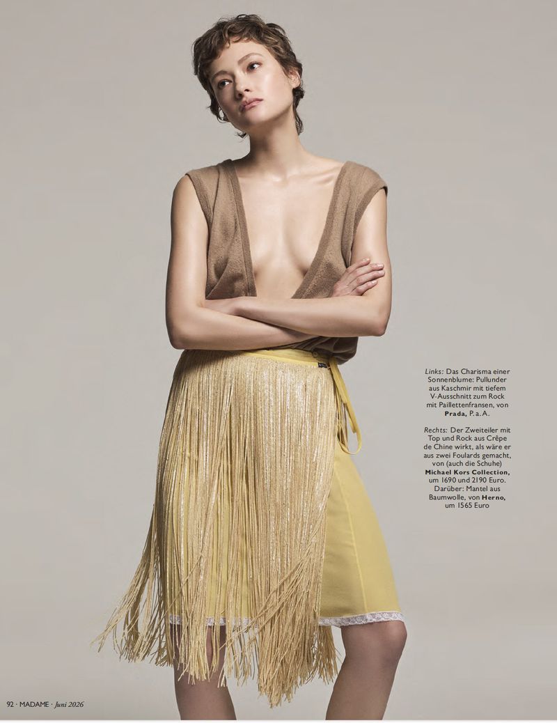 Model with short brown hair wearing a deep V-neck knit top and yellow fringed skirt against a neutral grey studio background