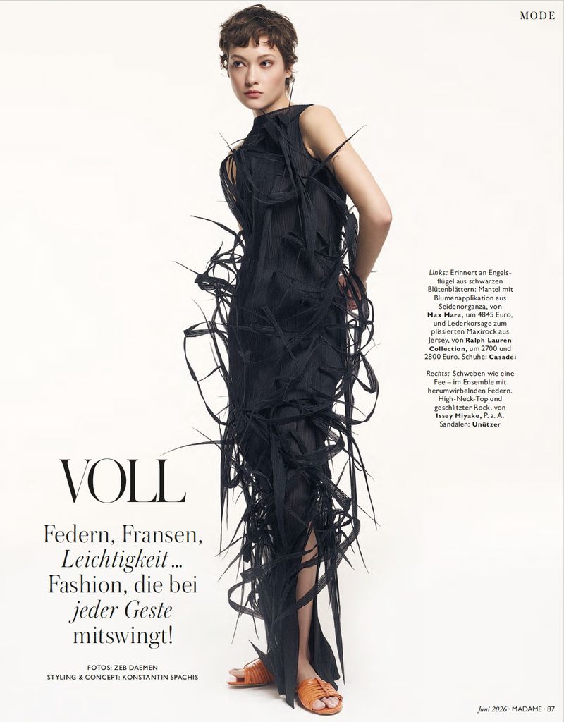 Model with short dark hair wearing a black fringed dress and orange sandals against a white studio background.
