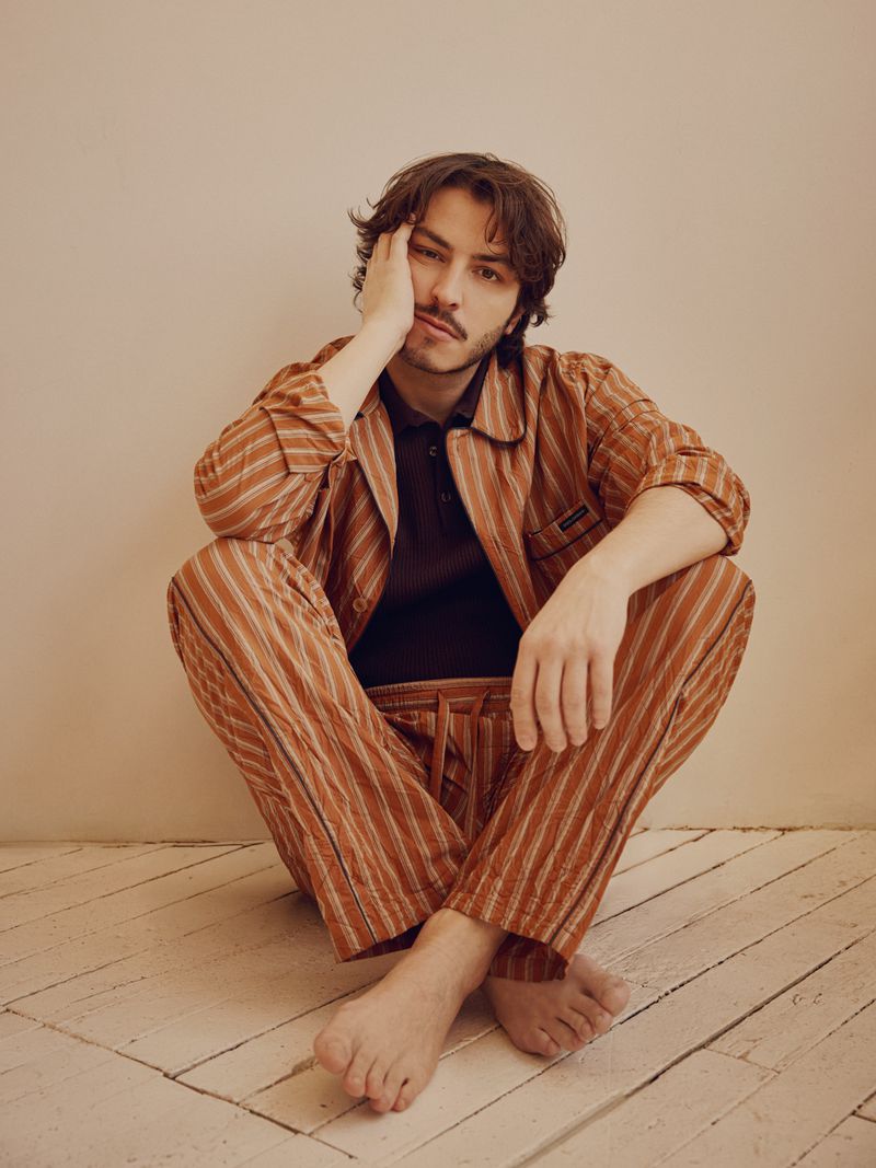 Boran Kuzum seated cross-legged on a pale wooden floor wearing a striped brown pajama-style shirt and matching trousers.