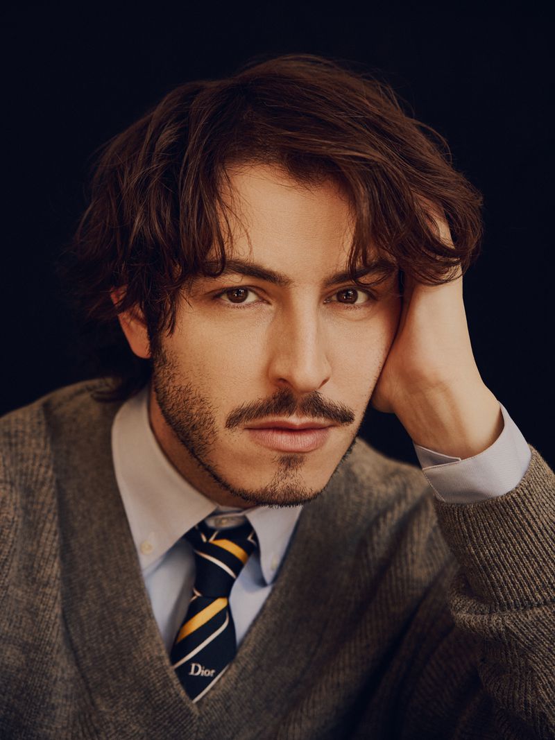 Portrait of actor Boran Kuzum wearing a grey sweater over a light blue collared shirt and striped tie against a dark background