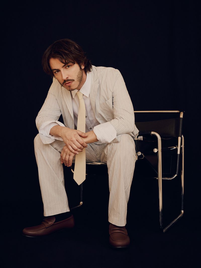 Actor Boran Kuzum seated on a black chair wearing a pinstripe suit and cream tie against a solid black background.