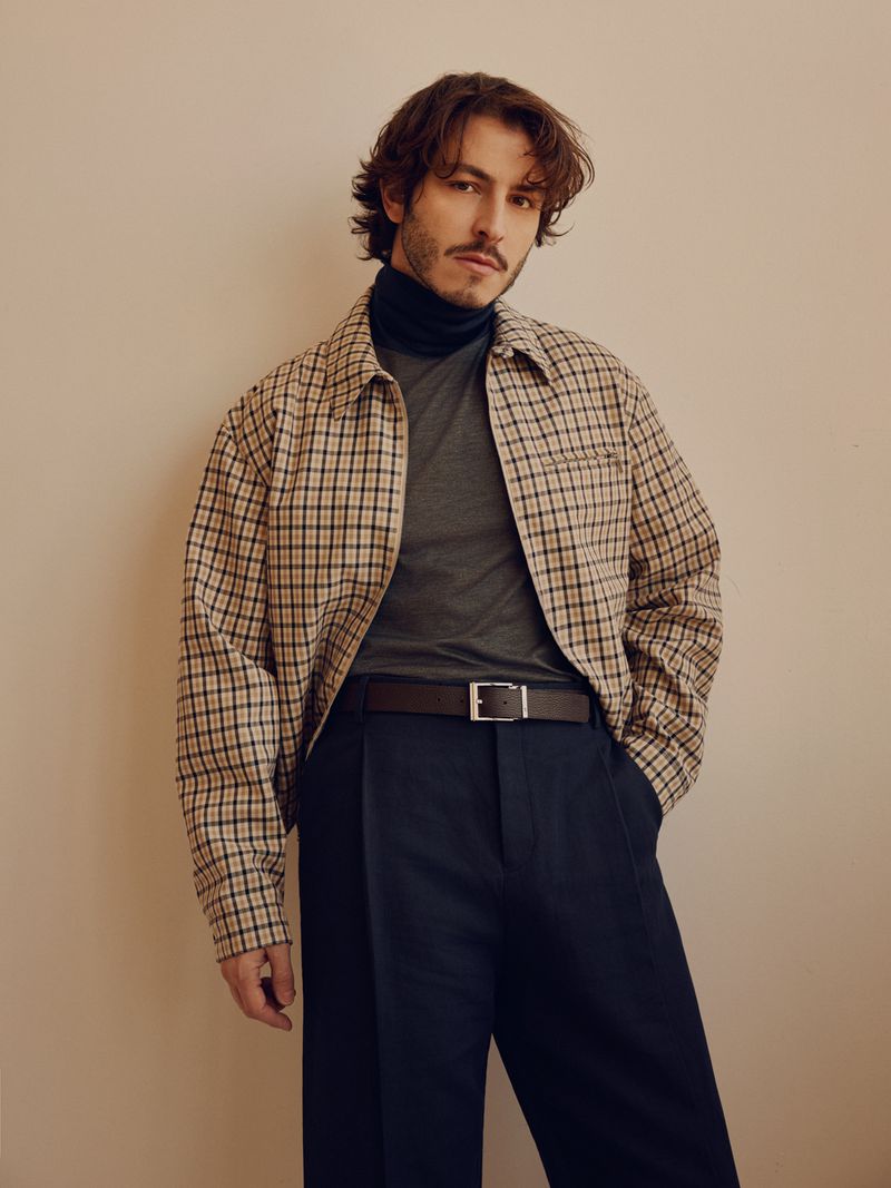 Man in a plaid jacket, dark shirt and trousers, posing indoors with a neutral backdrop, in a fashion portrait showcasing modern menswear.