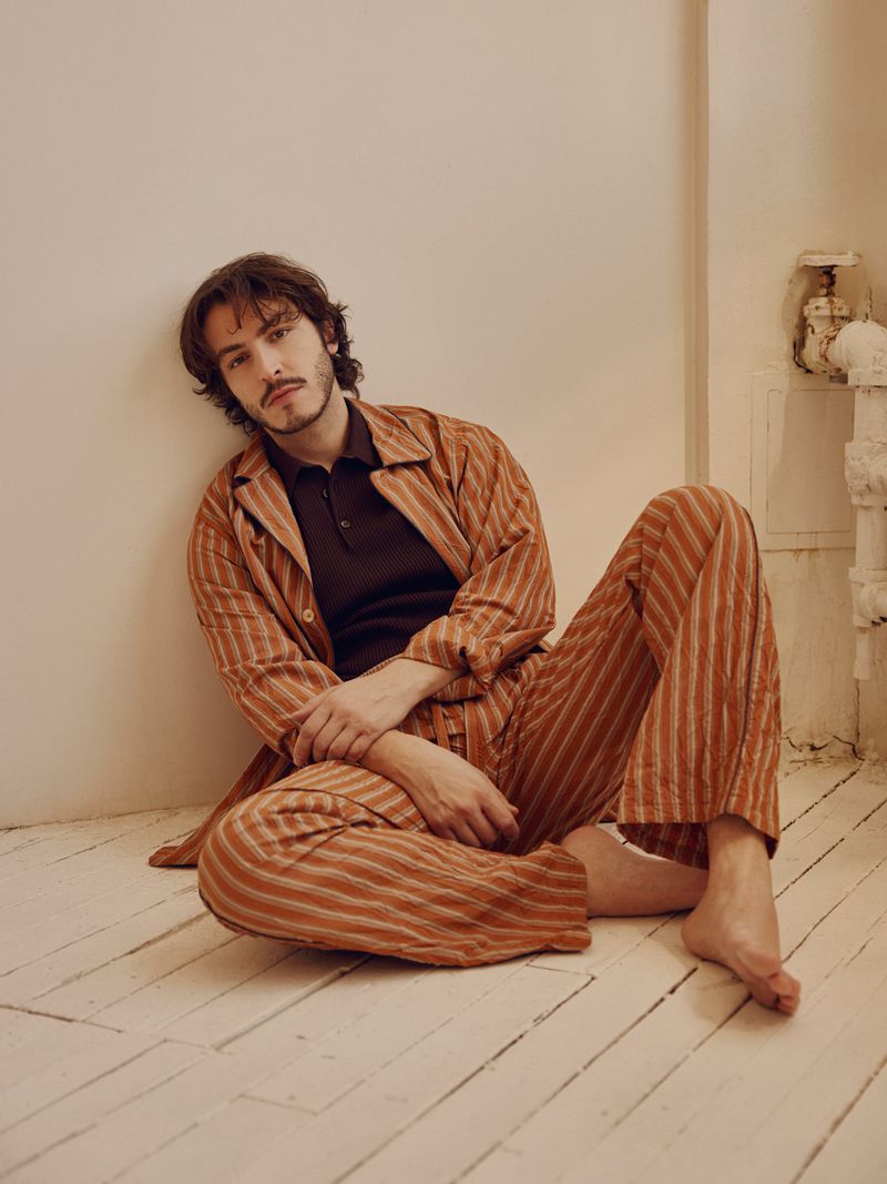 Actor Boran Kuzum seated on a white wooden floor leaning against a wall wearing a striped brown pajama-style set and dark polo shirt