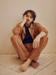 Boran Kuzum seated cross-legged on a pale wooden floor wearing a striped brown pajama-style shirt and matching trousers.