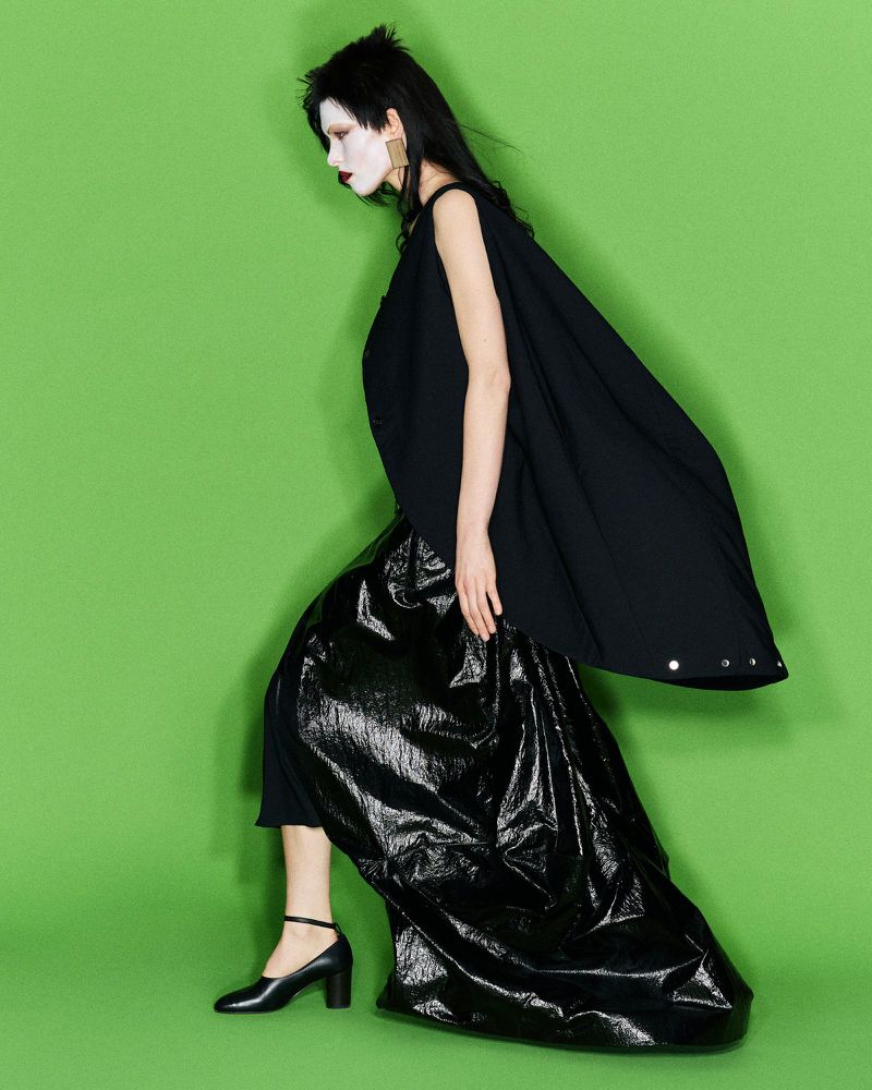 Model with dark hair wearing a flowing black cape and shiny black skirt against a vibrant green backdrop