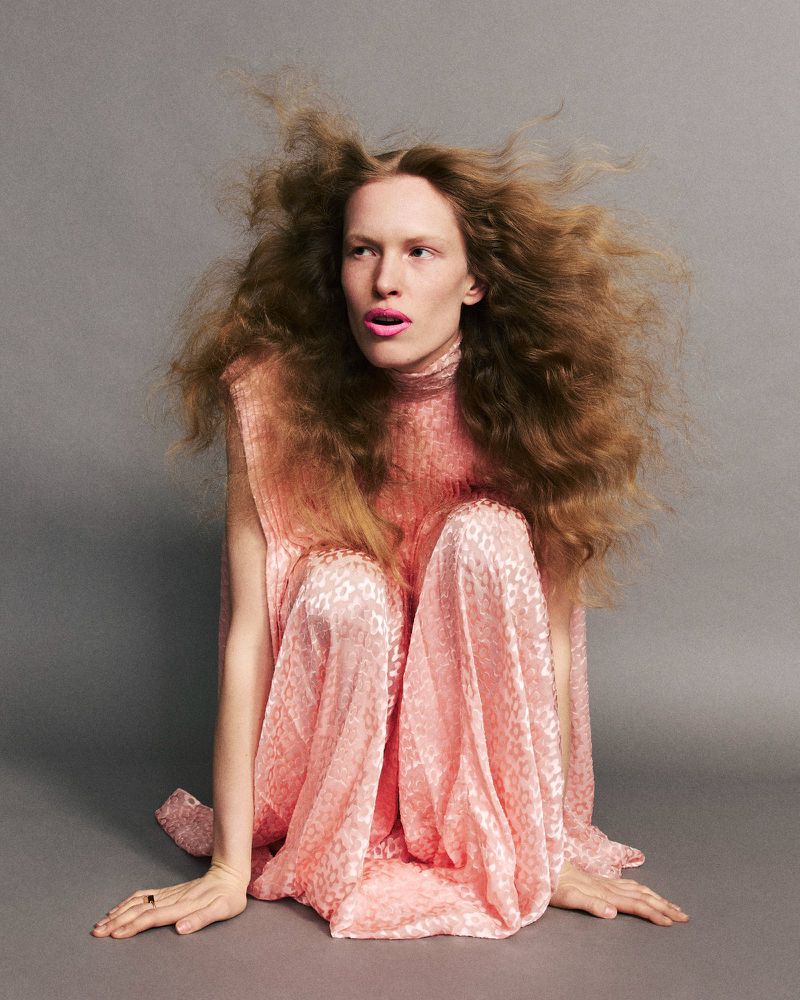 Model with voluminous hair wearing a pink patterned dress crouched against a grey seamless studio background.