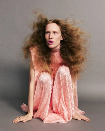 Model with voluminous hair wearing a pink patterned dress crouched against a grey seamless studio background.