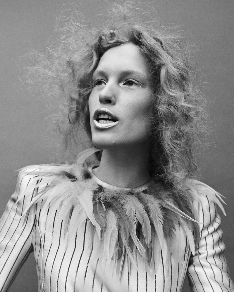 Model with textured curly hair and feathered collar wearing a striped jacket against a neutral backdrop.