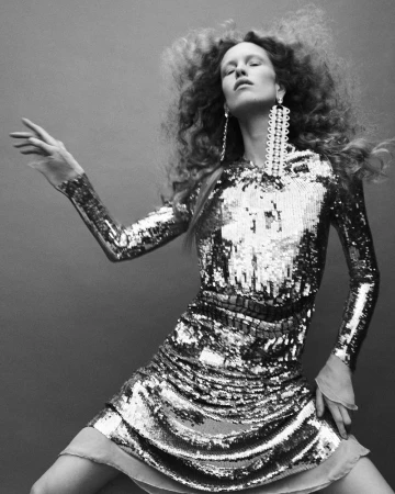 Model with voluminous curly hair wearing a sequined dress and long earrings against a neutral grey studio backdrop.