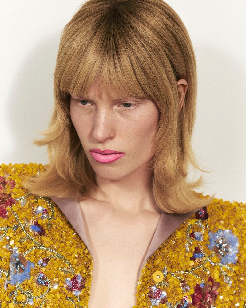 Close-up portrait of a model with blonde hair and bright pink lipstick wearing a heavily textured yellow sequin jacket against a plain background.