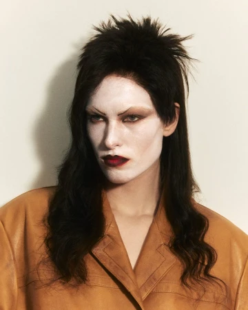 Model with dark layered hair and pale dramatic makeup wearing a tan jacket against a plain background