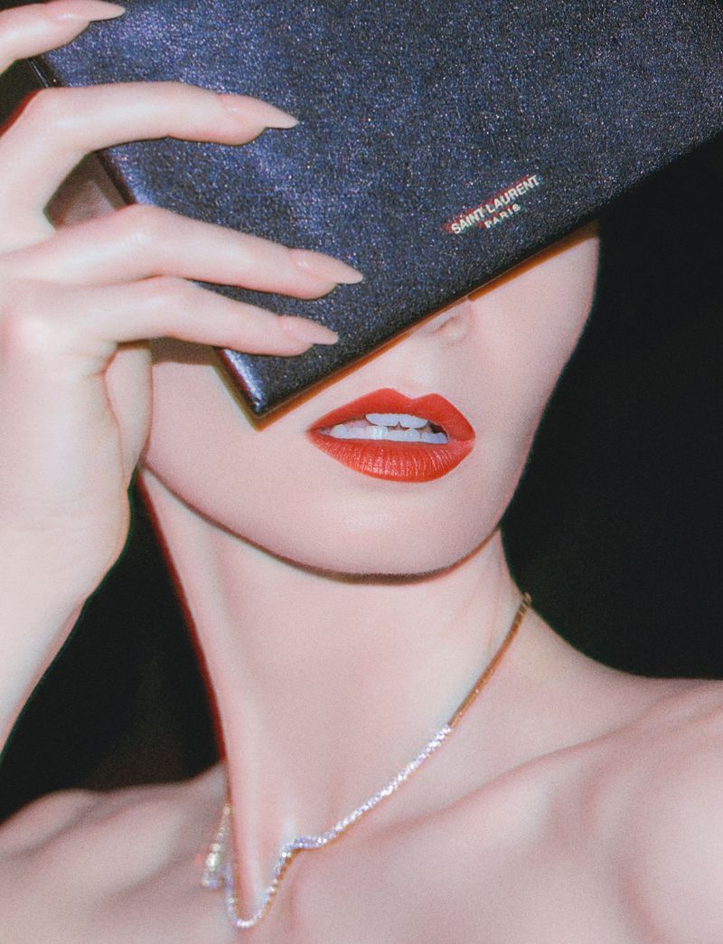 Close-up of a woman holding a dark clutch over her eyes, featuring red lipstick and a diamond necklace against a black background.