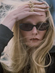 Close-up of a blonde woman wearing dark sunglasses with her hand across her forehead, displaying gold rings.
