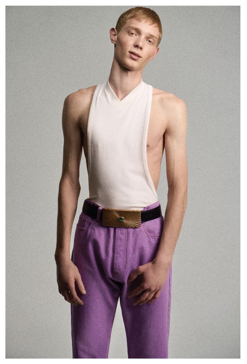 A young male model with short red hair wearing a sleeveless white top and purple trousers against a grey backdrop.