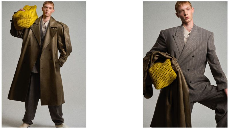 Male model wearing a grey pinstripe suit and leather coat holding a yellow woven bag against a neutral grey studio background