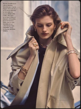 Model wearing a beige trench coat and gold jewellery adjusting her collar in an outdoor setting for Elle France