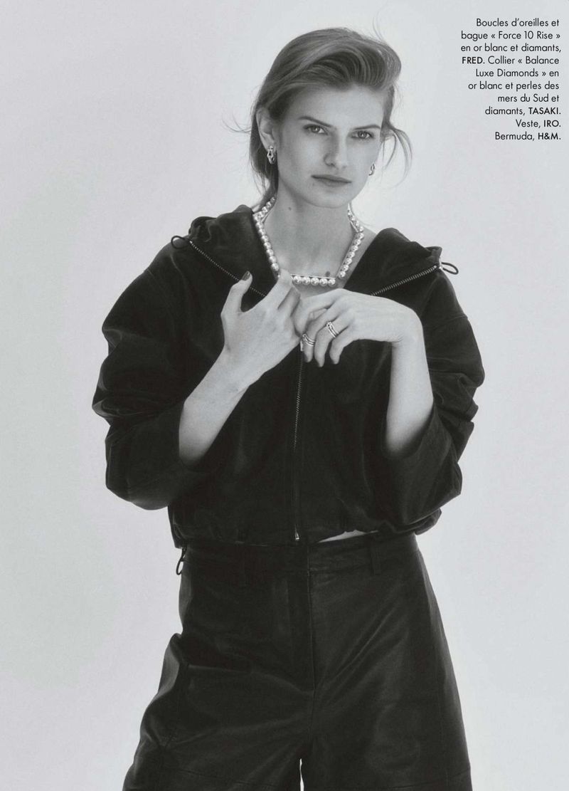 Model wearing a black zip-up jacket and pearl necklace against a seamless white background in black and white.