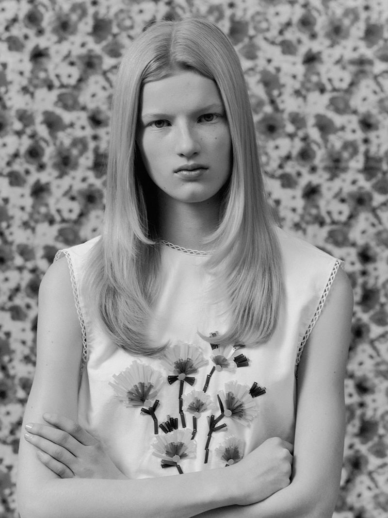 Monochrome portrait of a fair-skinned woman with blonde layered hair, wearing a white sleeveless top with floral embellishments, floral background.