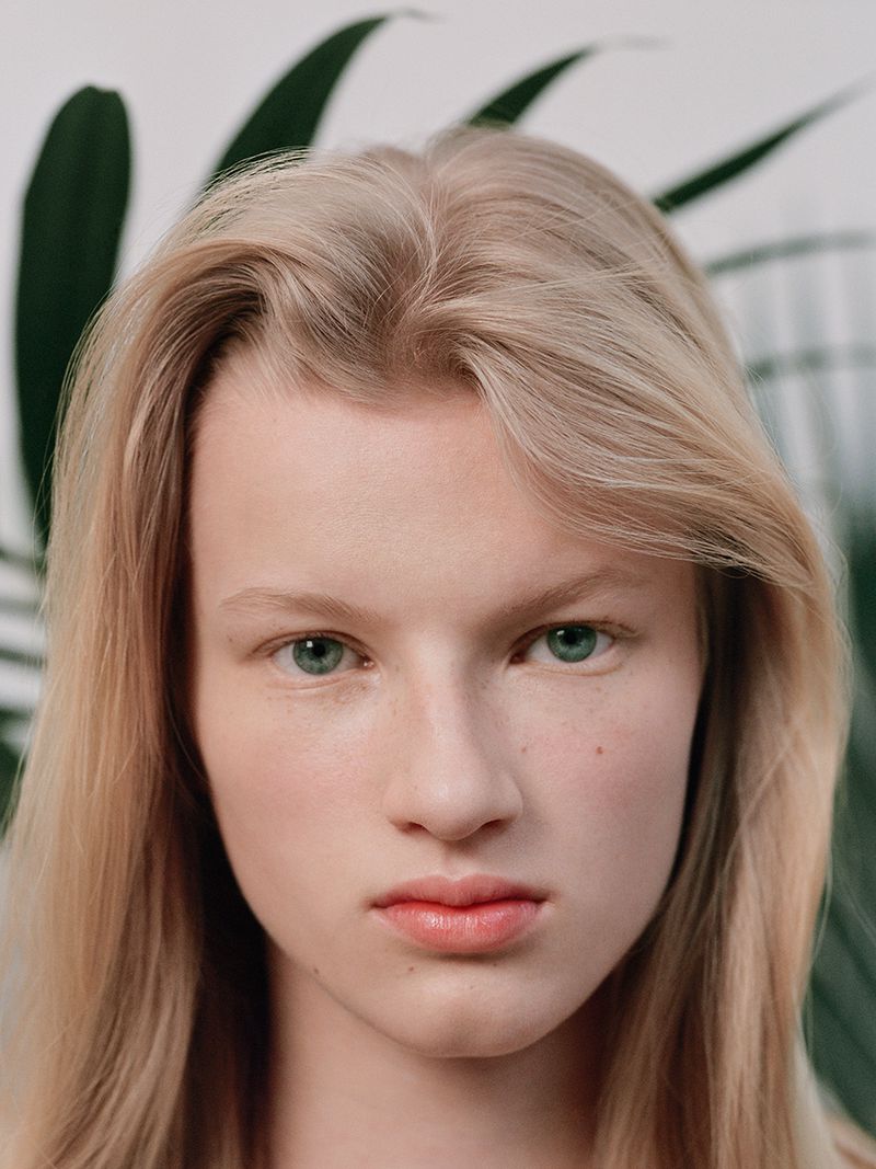 Close-up portrait of a fair-skinned adult female with long blonde hair, green eyes, and coral lips, posing in front of green tropical leaves.