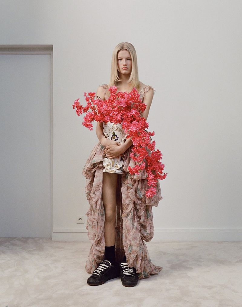Fashion photo of a blond model in an elaborate dress with a floral vase, posing in a white room with sneakers and socks.