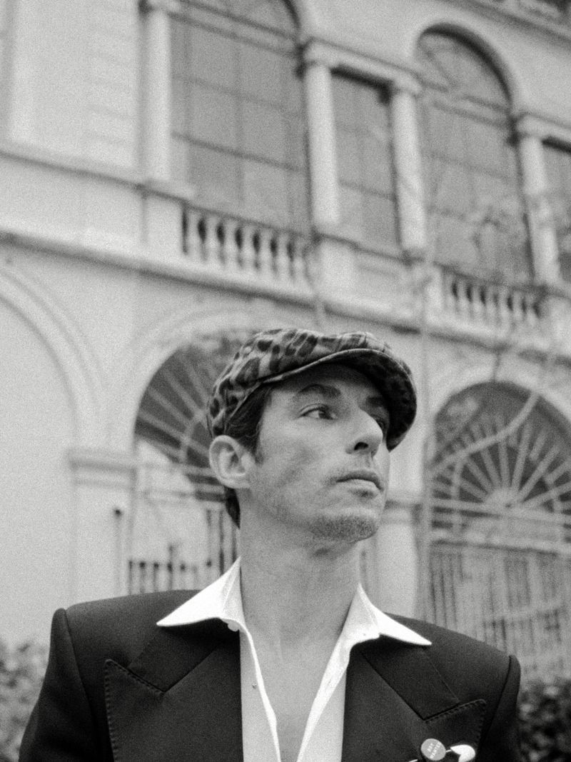 Portrait of actor Alberto Guerra wearing a patterned flat cap and dark blazer with a white shirt in front of a classical building.