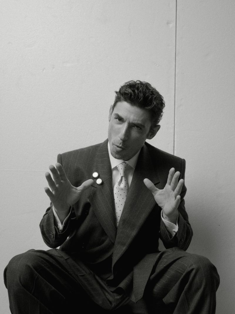 Monochromatic portrait of a man in a suit with an open-mouthed expression, gesturing with both hands in an indoor setting against a plain background.