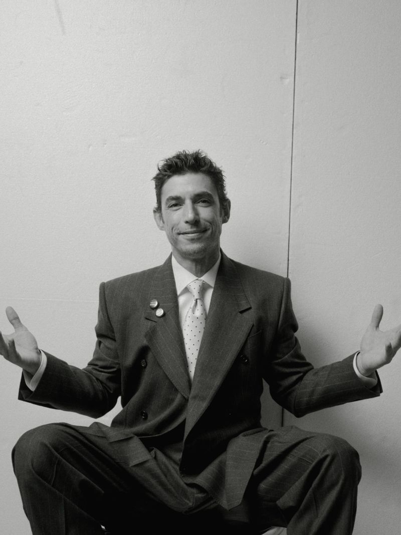 Portrait of actor Alberto Guerra wearing a patterned suit jacket and tie while gesturing outdoors.
