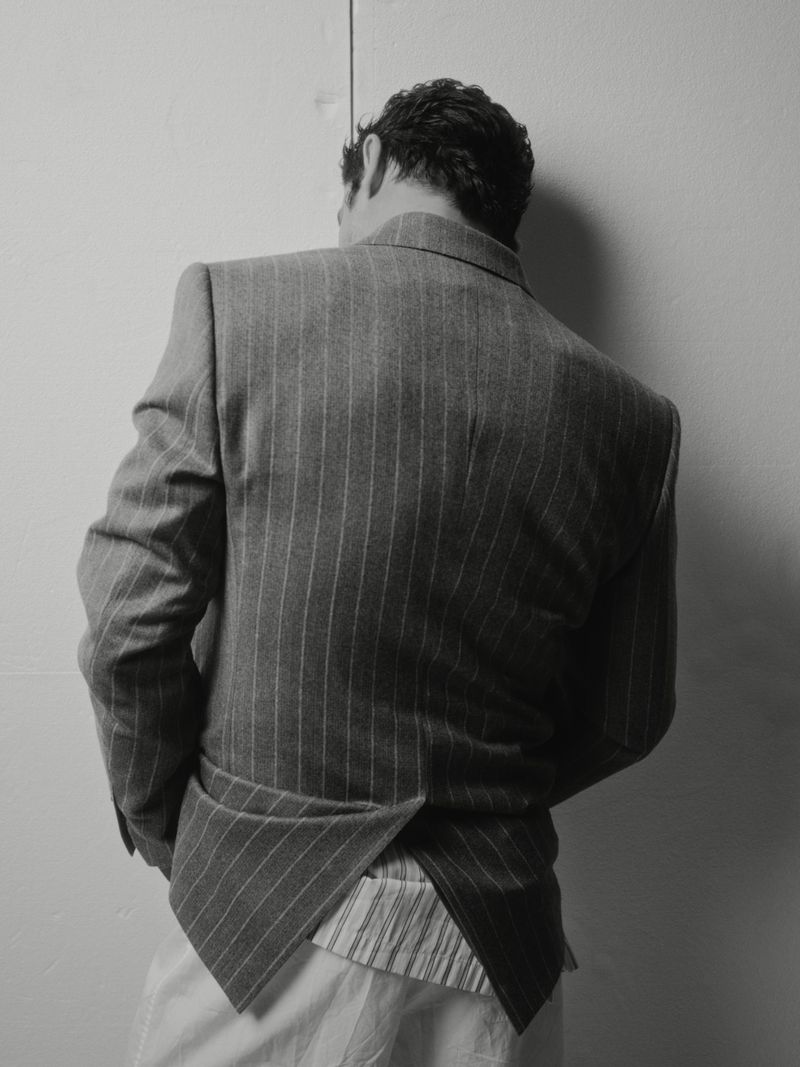 Man wearing a structured pinstripe suit jacket viewed from the back against a pale studio wall