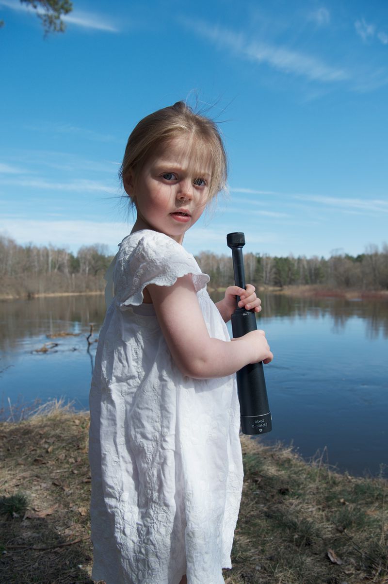 Young child in white dress holding black cylinder object by riverbank under blue sky