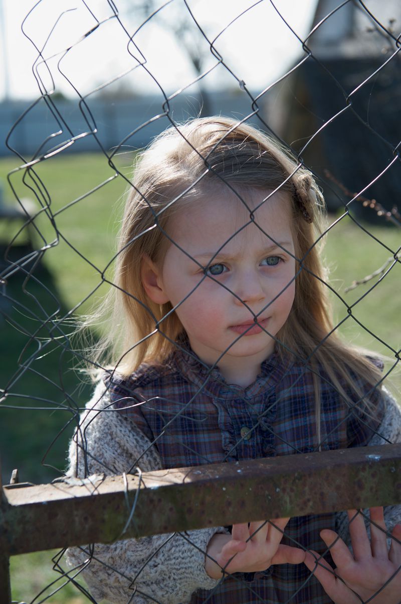 Young child with blonde hair standing behind a wire fence outdoors in natural light.
