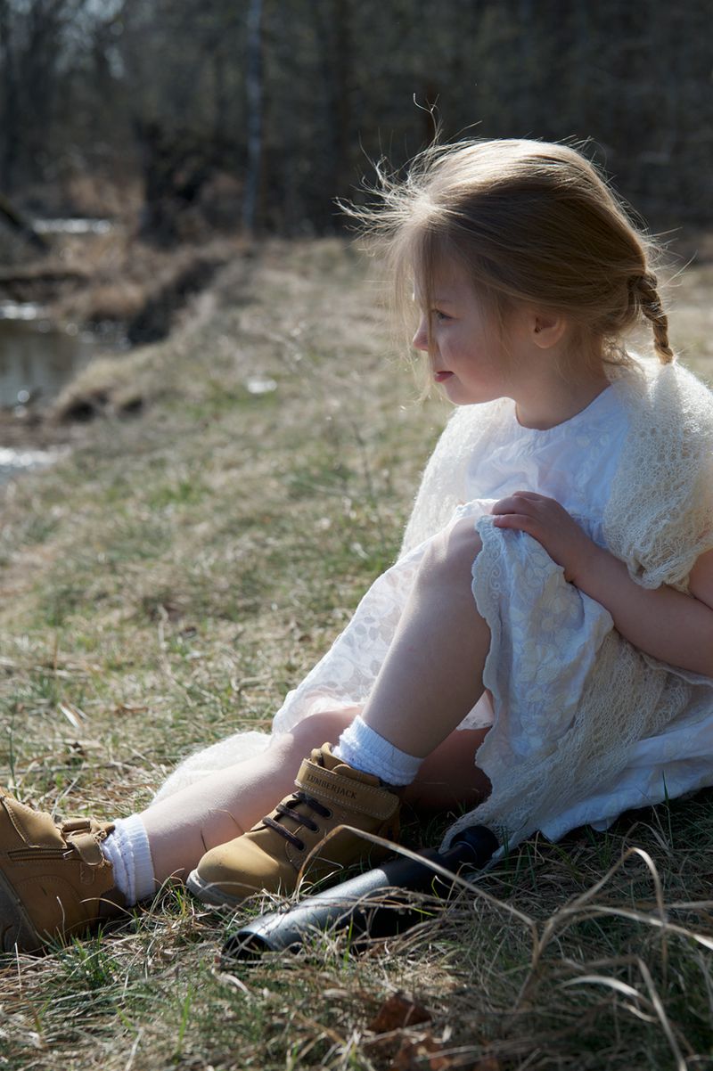 Young child in white dress and brown boots seated outdoors on grassy ground near a woodland edge