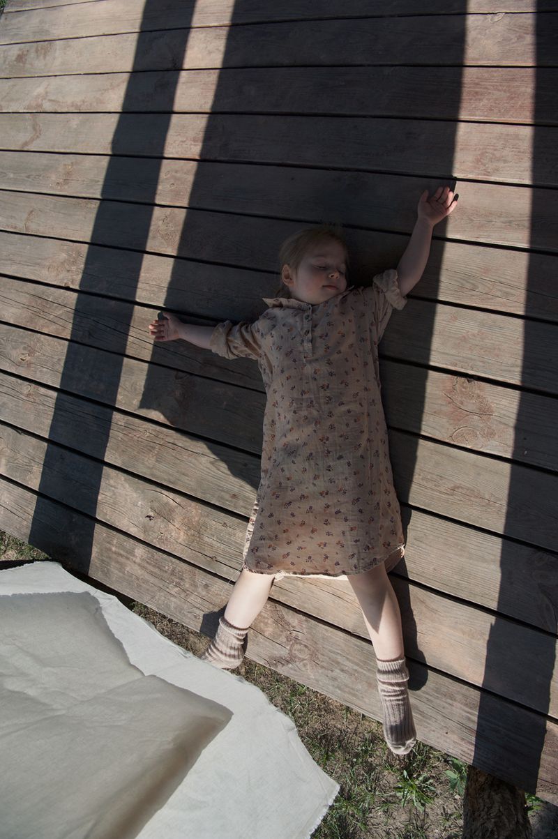 Child lying flat on a wooden deck outdoors wearing a patterned dress and ribbed socks with long cast shadows.