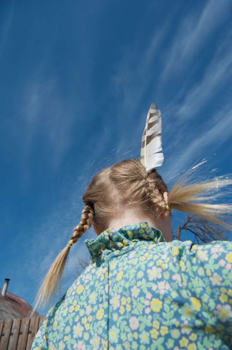 Rear view of a young child with blonde hair in pigtails wearing a patterned dress with a feather tucked into her hair against a clear blue sky