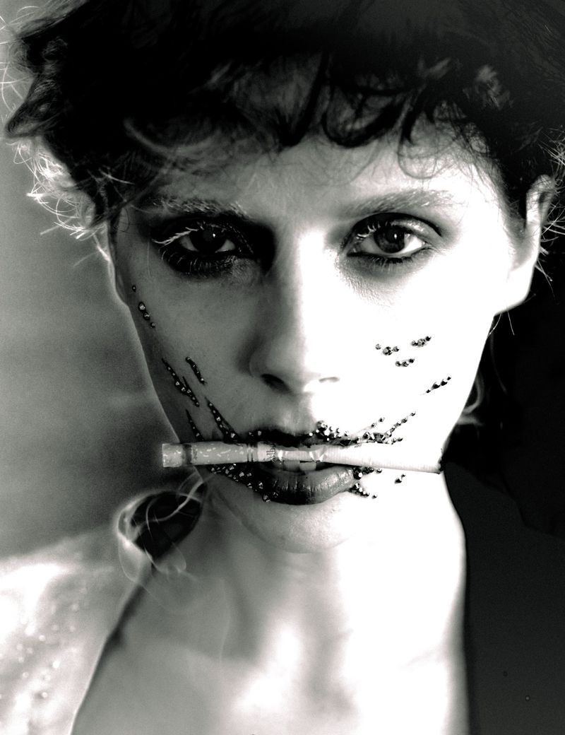Close-up monochrome portrait of a woman with dark curly hair holding a cigarette in her mouth, featuring textured face details.
