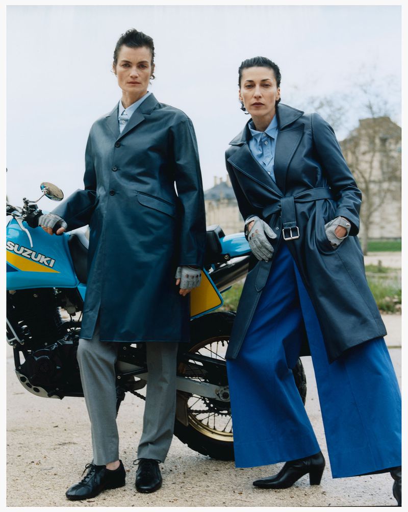 Two female models in dark blue coats standing outdoors next to a blue motorcycle under an overcast sky.