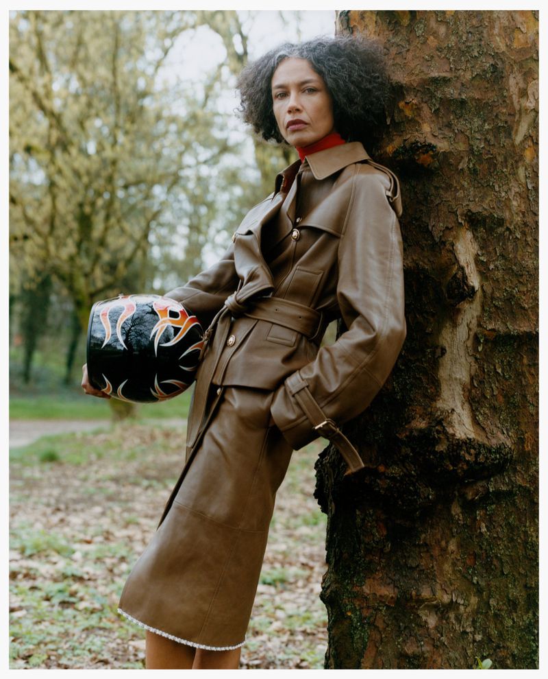 Model leaning against a tree wearing a brown leather coat and holding a patterned helmet in a woodland setting.