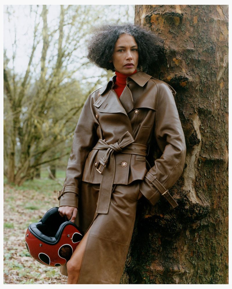 Model leaning against a tree wearing a brown leather coat and holding a red helmet in a woodland setting.