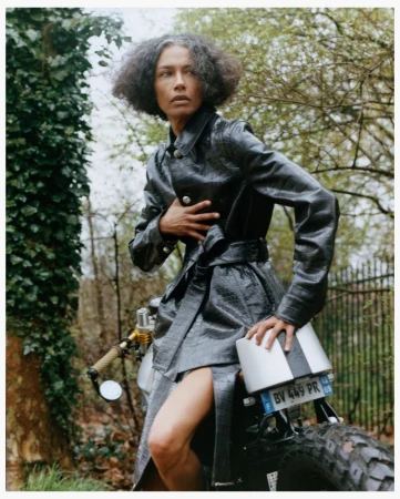 Model in a black textured coat seated on a motorcycle outdoors near a tree and fence