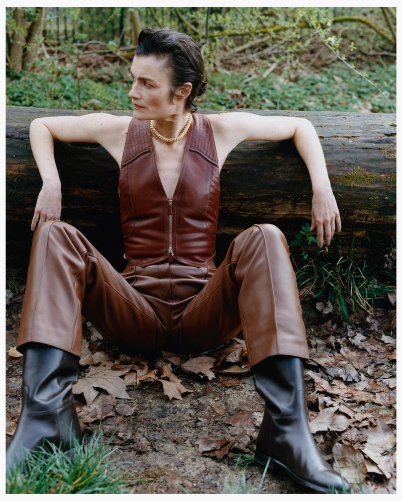 Model wearing brown leather vest and trousers sitting against a fallen log in a forest