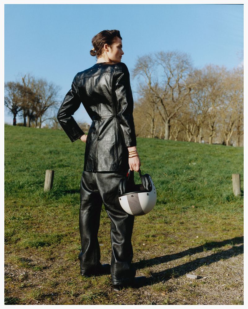 Model in a black leather jacket and trousers holding a motorcycle helmet outdoors on a grassy hill under a blue sky.