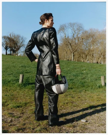 Model in a black leather jacket and trousers holding a motorcycle helmet outdoors on a grassy hill under a blue sky.