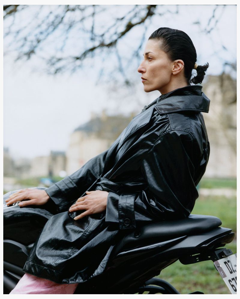 Model in black leather coat seated on a motorcycle outdoors with blurred building exterior in the background