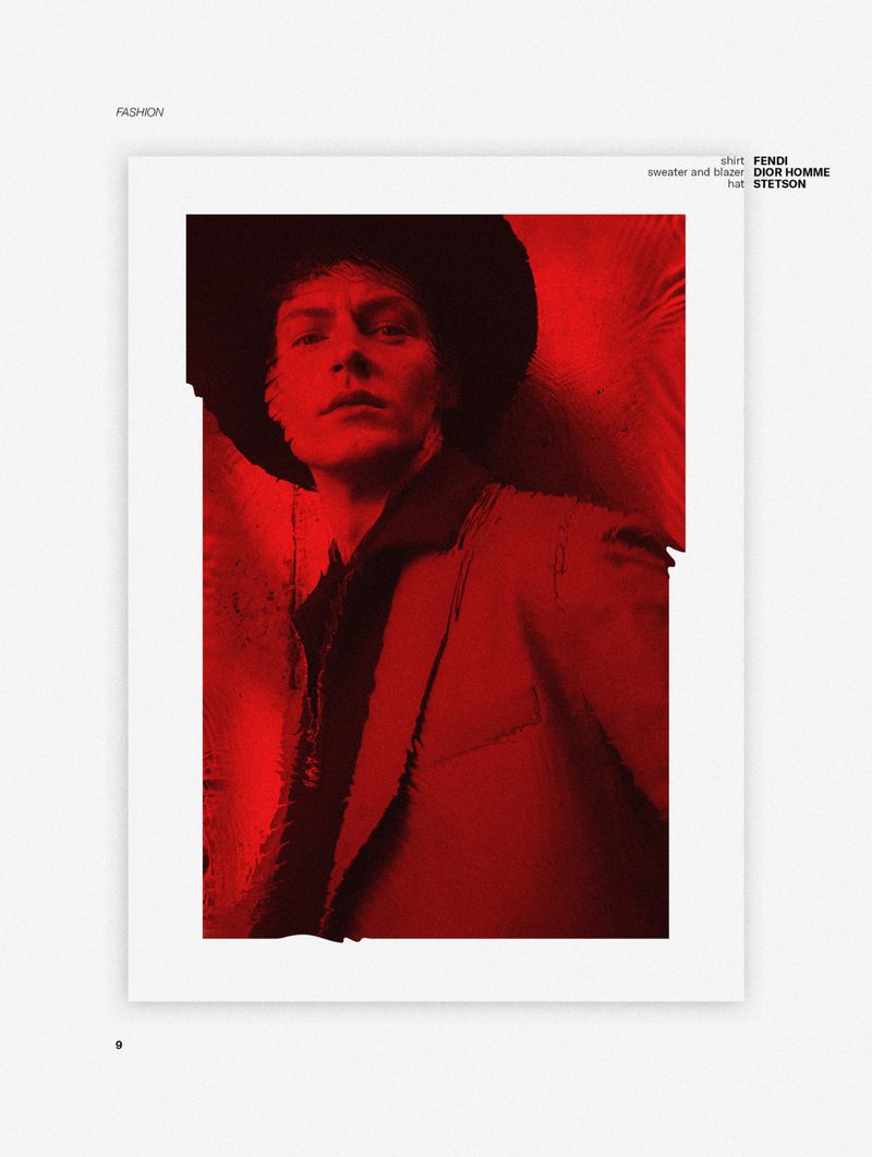 Model in wide-brim hat wearing layered blazer and dark shirt under intense red lighting in fashion editorial portrait.