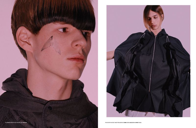 Close-up portrait of a male model in a dark hooded jacket with a distinct bowl cut hairstyle against a pale background