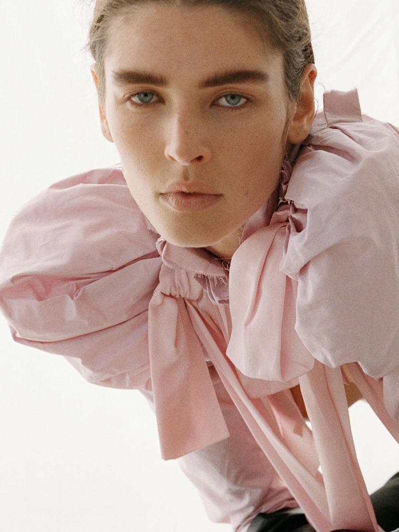 Close-up of a model wearing a large, light pink puffed-sleeve blouse with bows, posing in a photo studio with bright, neutral lighting.