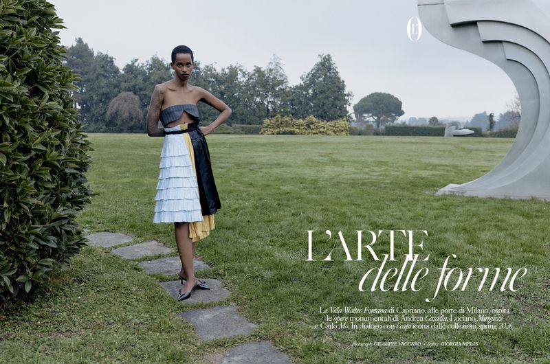 Model standing on stone path in a green park wearing a strapless top and pleated white skirt for editorial shoot