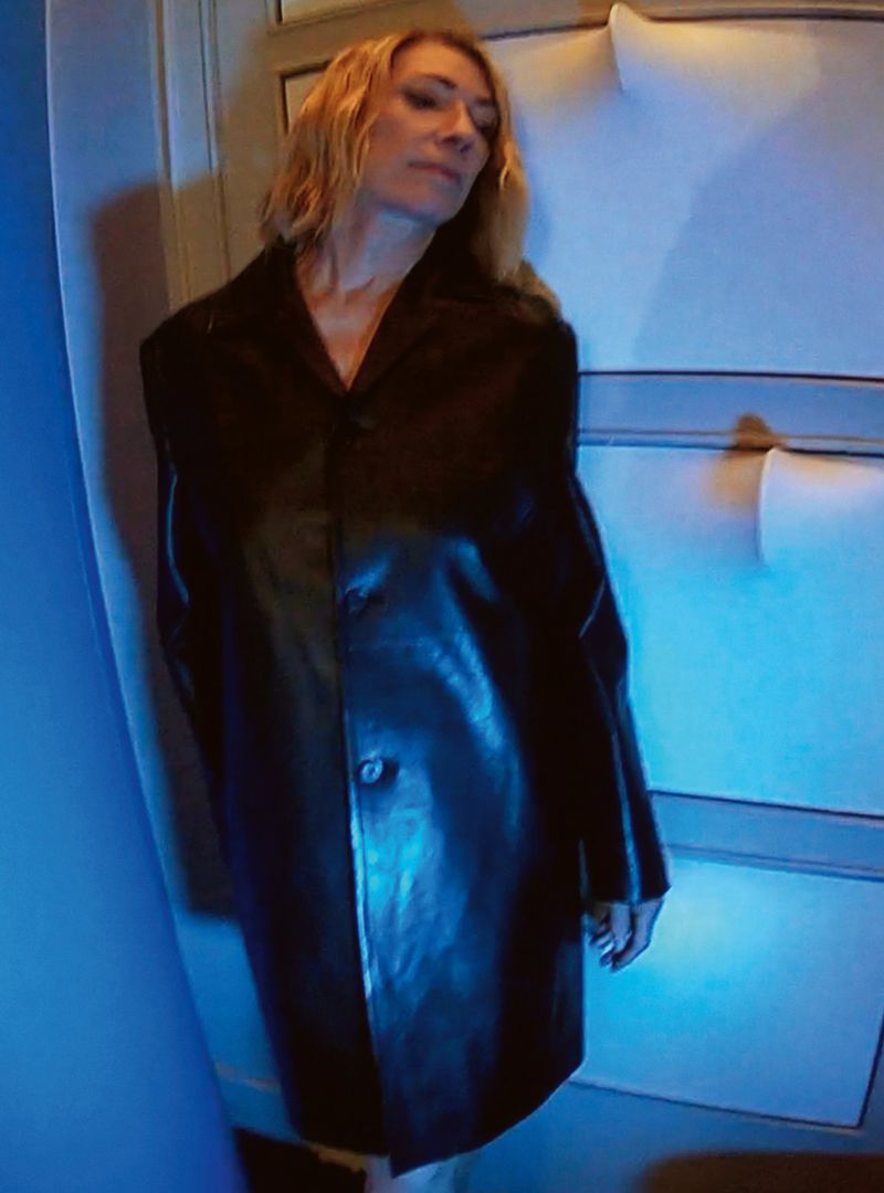Indoor portrait of a blond woman in a long black coat, with a neutral expression, standing near a white wall with blue light.