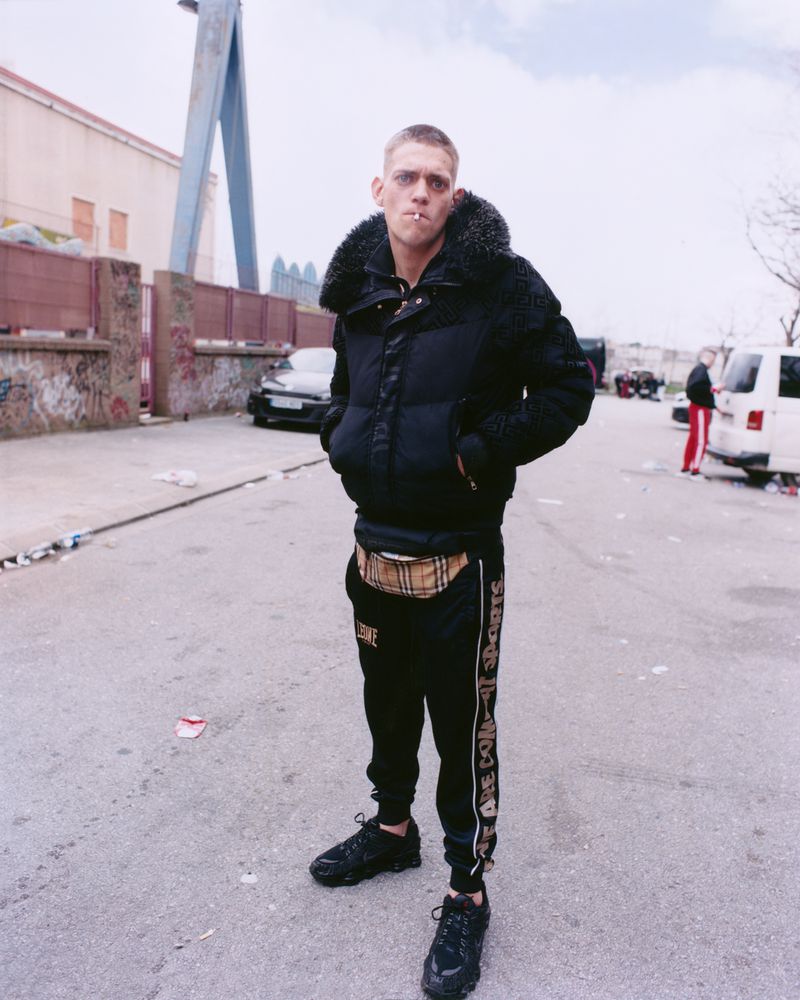 Adult male in puffer jacket and tracksuit poses on urban street with cars and buildings under overcast sky in full-length shot.
