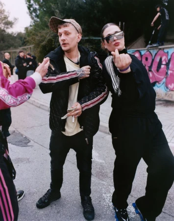 Two people posing outdoors on a street, with a man in a patterned jacket and a woman in sunglasses making a hand gesture.